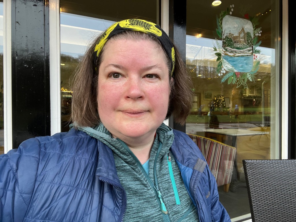 A selfie of me showing Head and shoulders. I have a slightly flushed face from running, have my hair back using a yellow and black ruff and am wearing a green running jacket under a blue winter jacket